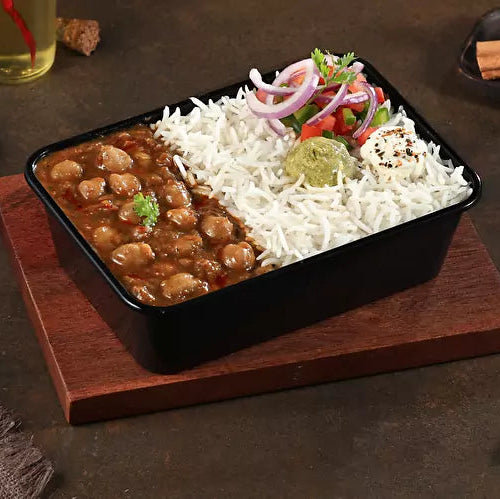 Black rectangular container with food on a wooden board, surrounded by spices and a wooden spoon on a dark surface.